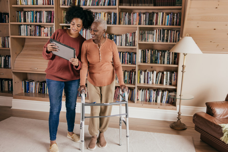 A woman and her mother, who is leaning on a walker, stand in a living room full of books.