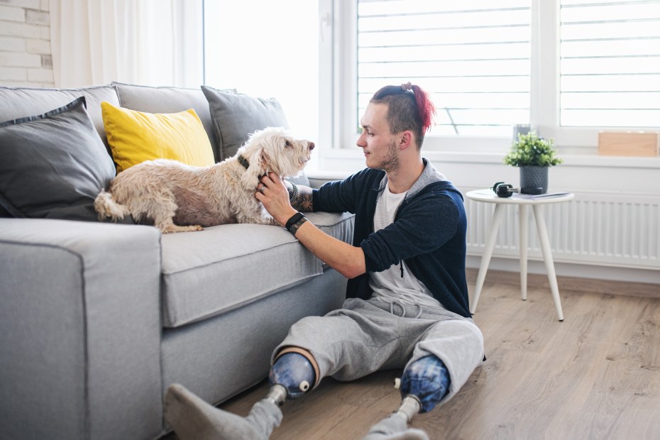 A man with prosethic legs plays with a small dog on a sofa.