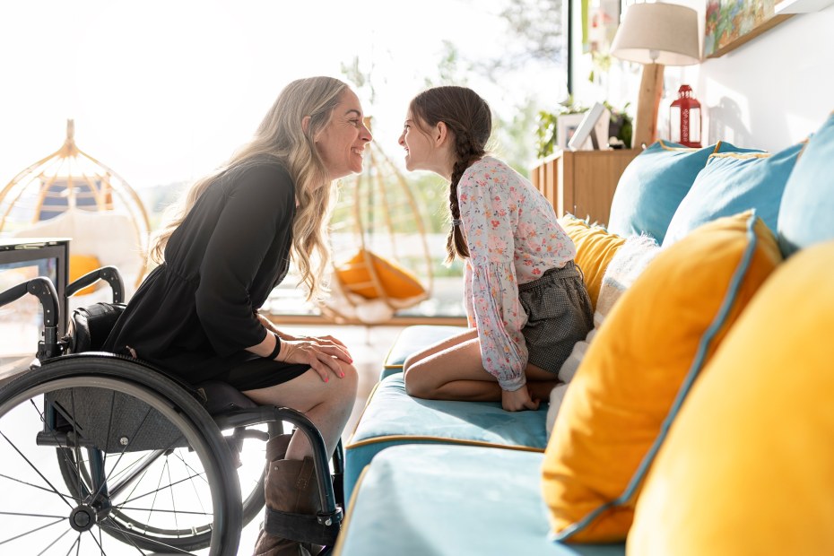 A woman in a wheelchair smiles at a girl perched on a sofa.