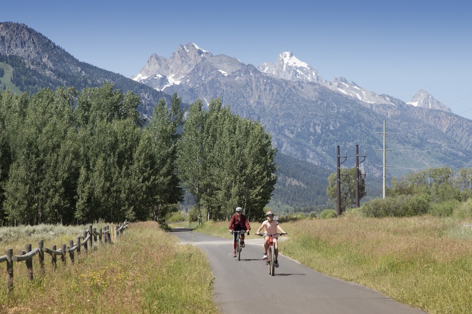 A parent and child bike on a paved trail with the Teton mountains in the background.