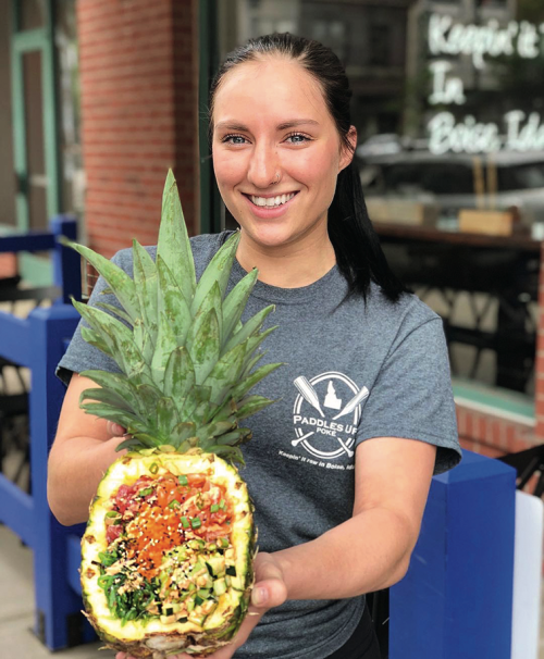 a pineapple half filled with poke ingredients held by a young woman