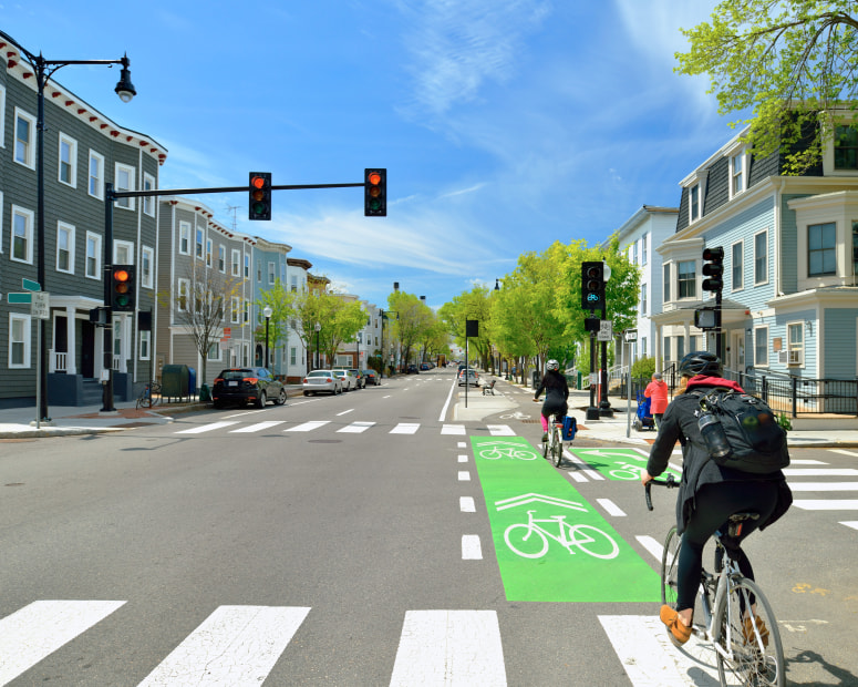 Cyclists go thorugh an intersection into a protected bike lane between parking lane and sidewalk on city street.