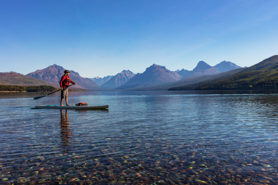 A person standup paddleboards on Lake McDonald in Glacier National Park, Montana on a clear day.