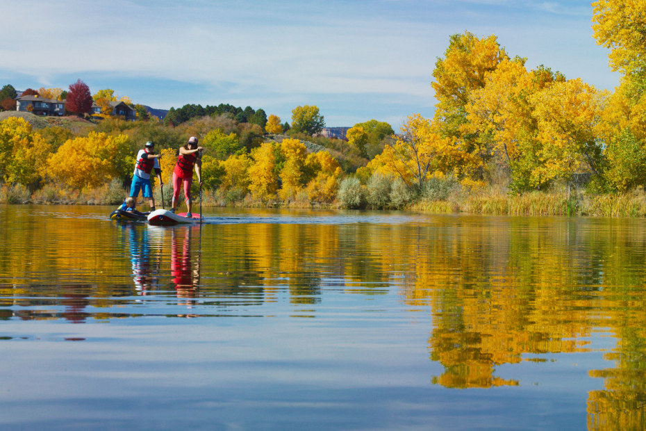 A couple paddleboard on a section of the Colorado River with fall colors behind them.