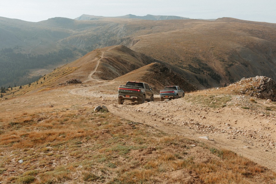 Two Rivian R1T drive downhill on a rocky, dirt trail.