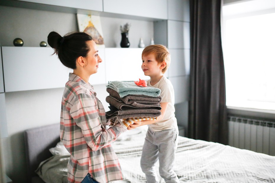 A mom hands her son a stack of towels with a bath toy on top.