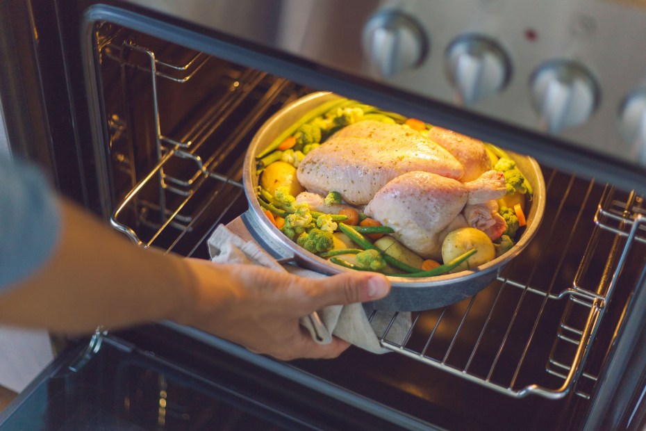 A home cook puts a chicken surrounded by vegetables into a hot oven.