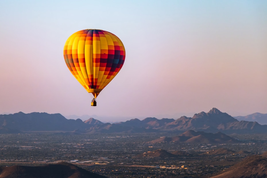 An orange, yellow, and brown hot balloon hovers over Phoenix, Arizona at sunrise.