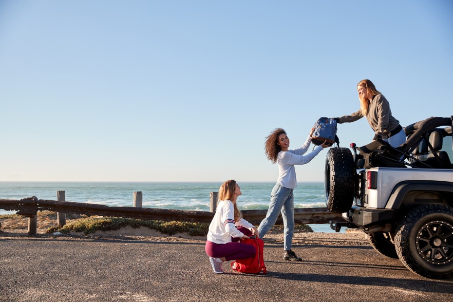 A group of friends unload their backpacks from the back of a Jeep parked by the ocean.