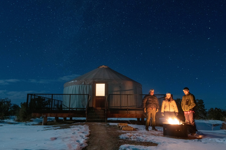 A group of campers stand around a campfire in winter with the Dead Horse State Park’s Wingate Campground yurt in the background.