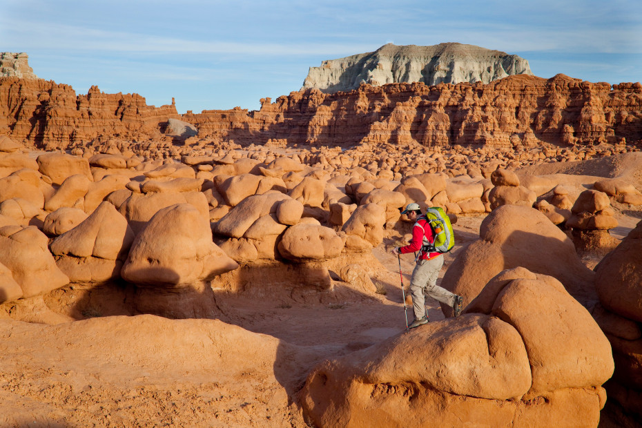 A hiker in a red jacket and a green backpack hikes through sandstone hoodoos in Goblin Valley State Park, Utah.