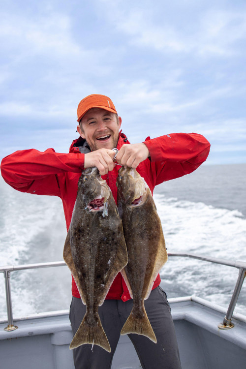A smiling fisherman holds two halibut caught on a deep sea fishing tour.