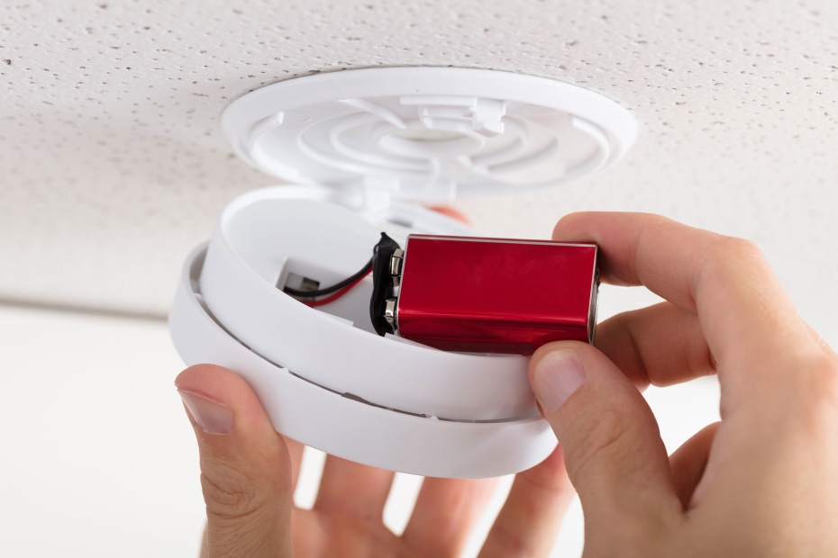 A person installs a new red battery into a smoke alarm on the ceiling.