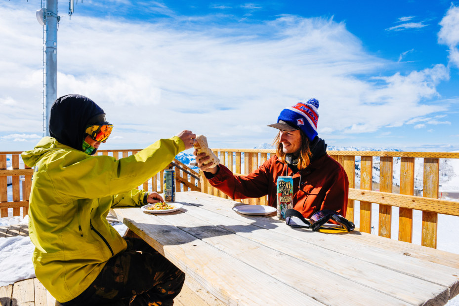 Two snowboarders cheers their burritos at a picnic table atop Powder Mountain in Utah.
