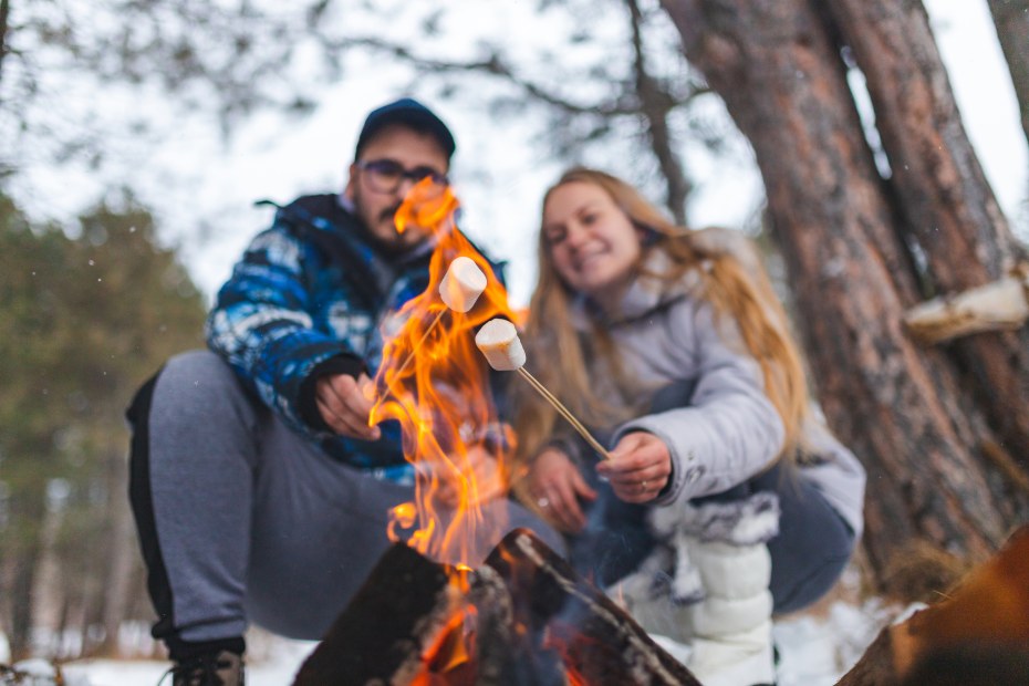 A couple roast marshmallows over the campfire with snow on the ground.