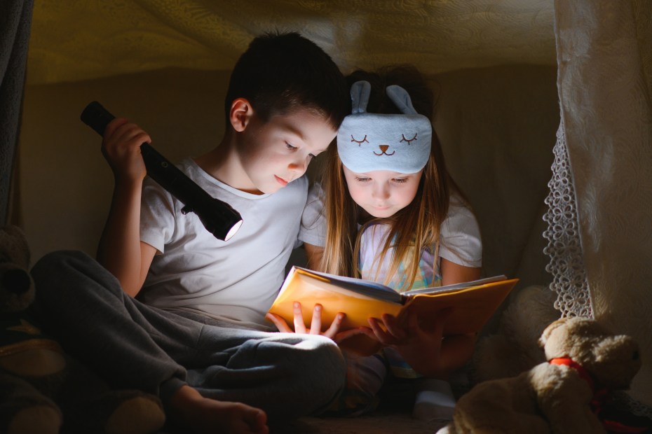 A brother and sister read a book by flashlight inside a blanket fort.