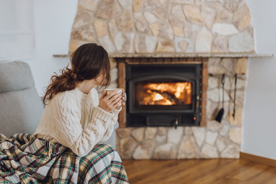 A woman wrapped in a plaid blanket and cream sweater sits in front of a gas fireplace.