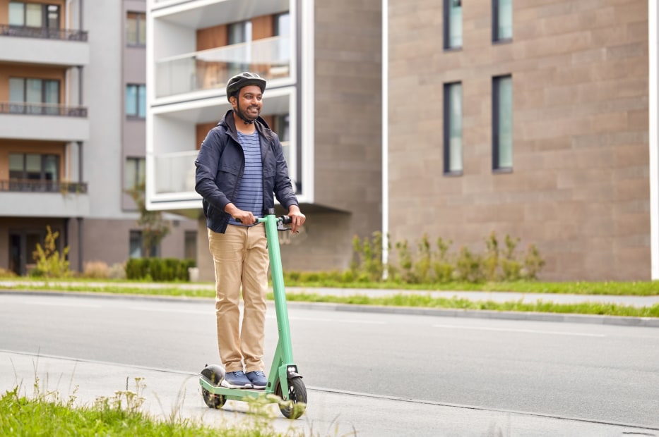 A man rides a green electric scooter with apartments in the background.