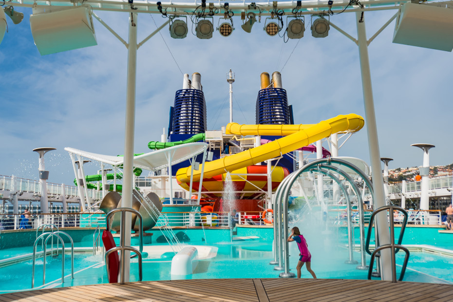 A young girl plays in the splash zone on a cruise ship with a waterslide in the background.