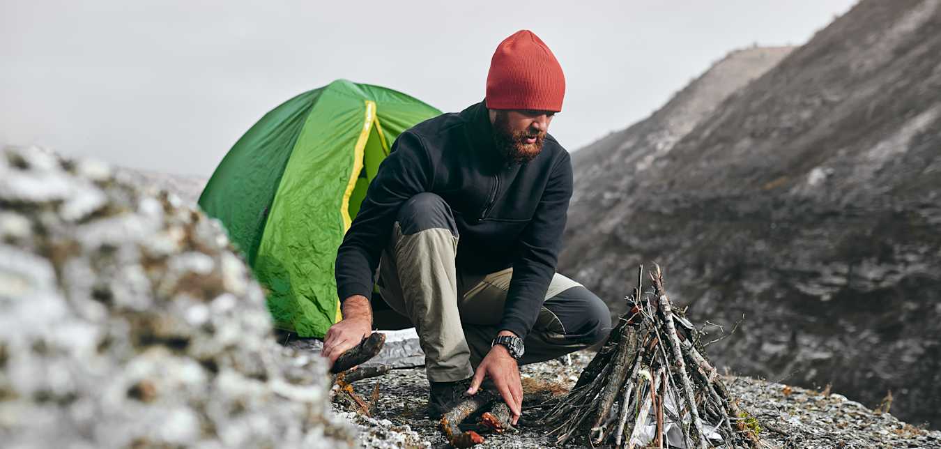 a hiker in a red beanie builds a campfire in front of a green tent