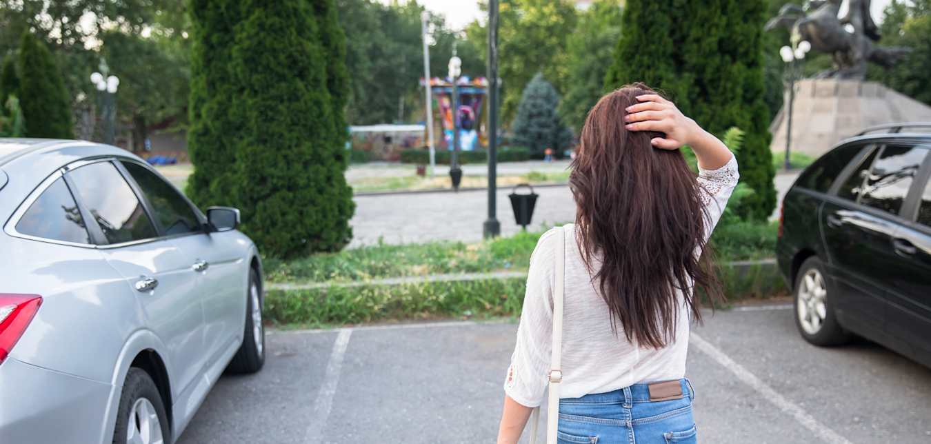 A woman with brown hair stands looking shocked in front of an empty parking space where her car should be.