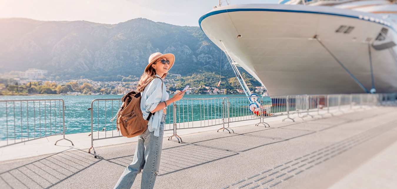 A young woman with a backpack smiles while walking towards a cruise ship