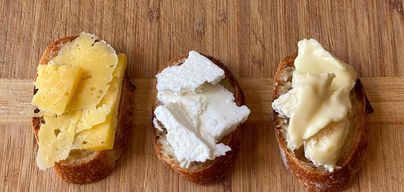 Three types of cheese top three slices of rustic bread on a wooden board.