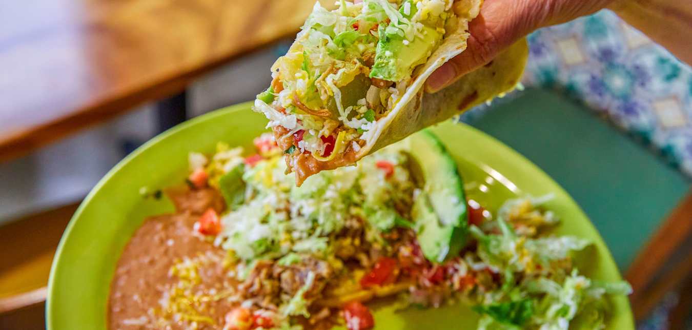 A hand holds a stuffed soft taco over a traditional Mexican combo plate. 