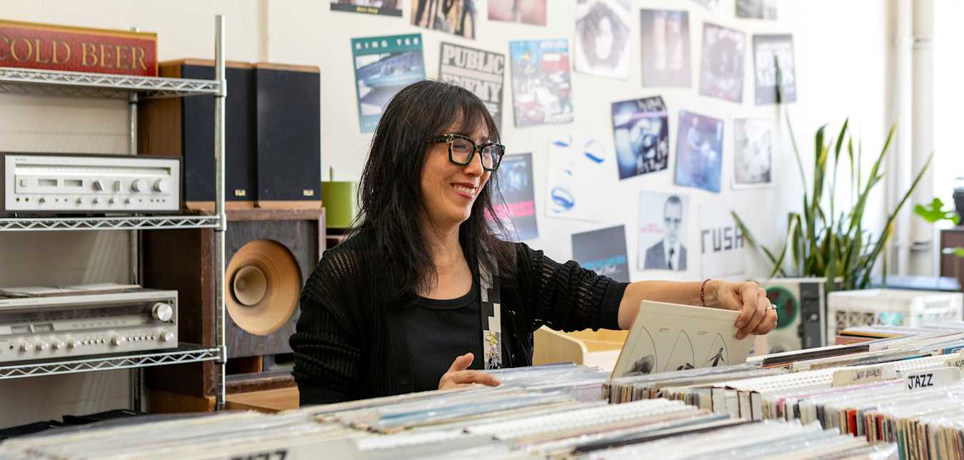 a woman with black hair and glasses smiles as she looks through records in a record store