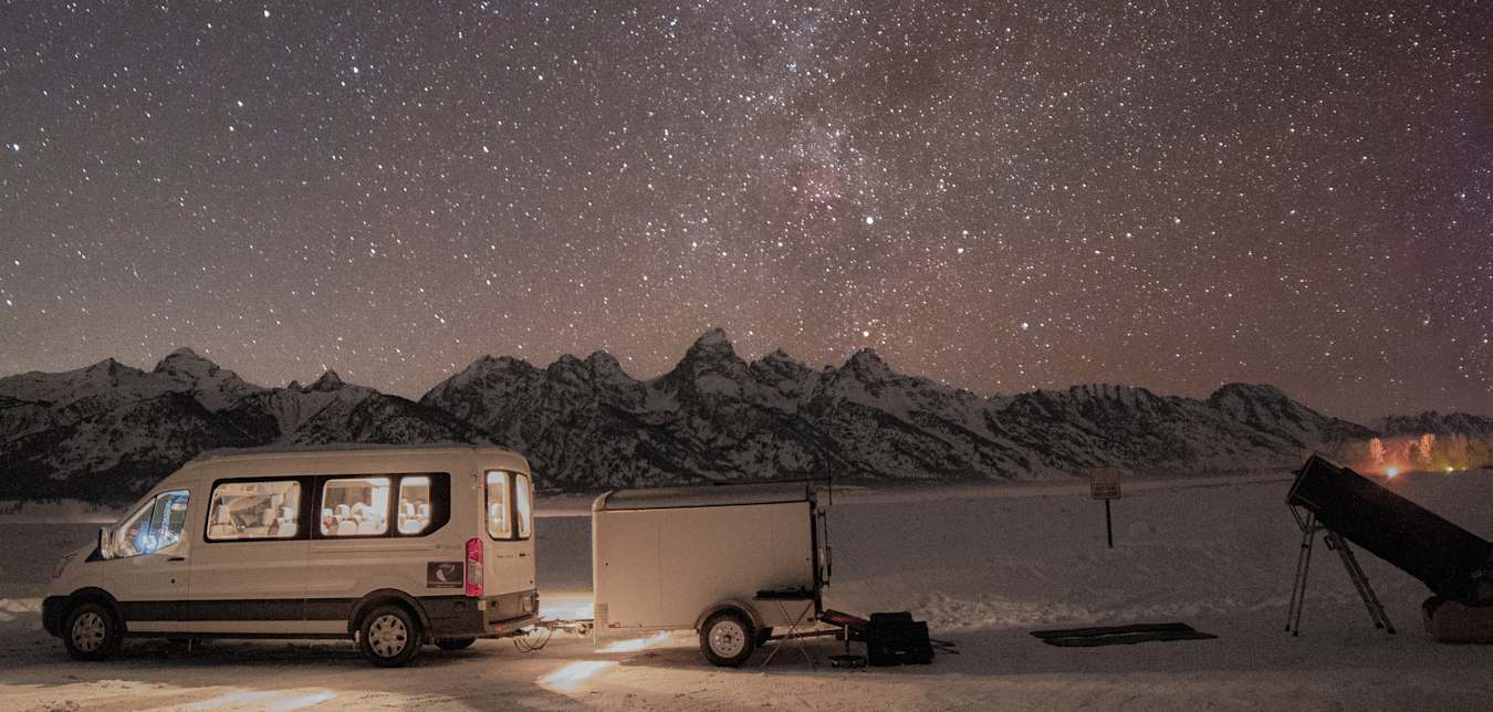 a van is parked near a telescope with a brilliant starry sky and mountains in the distance