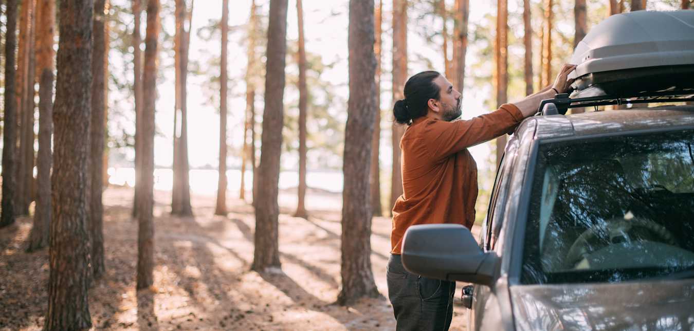 A person opens a roof-top storage box on their SUV parked in forest.