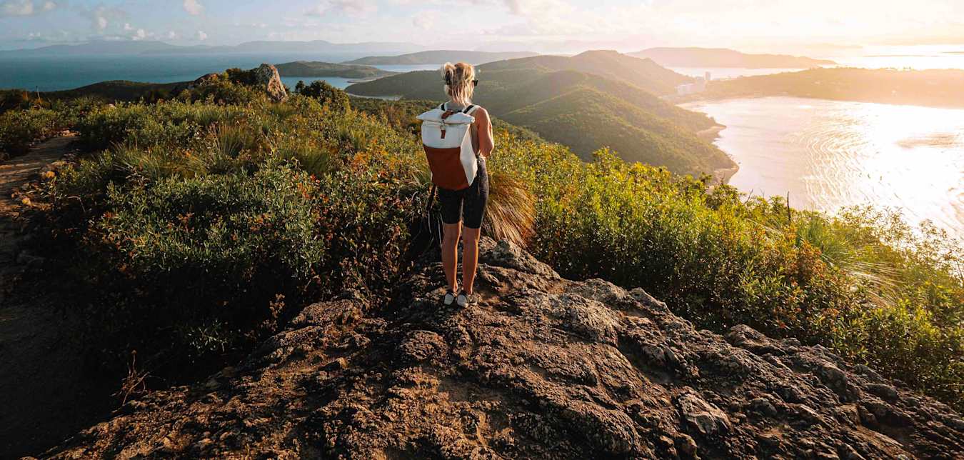 a woman searing shorts and a backpack stands on top of a mountain looking out at the ocean in the sun