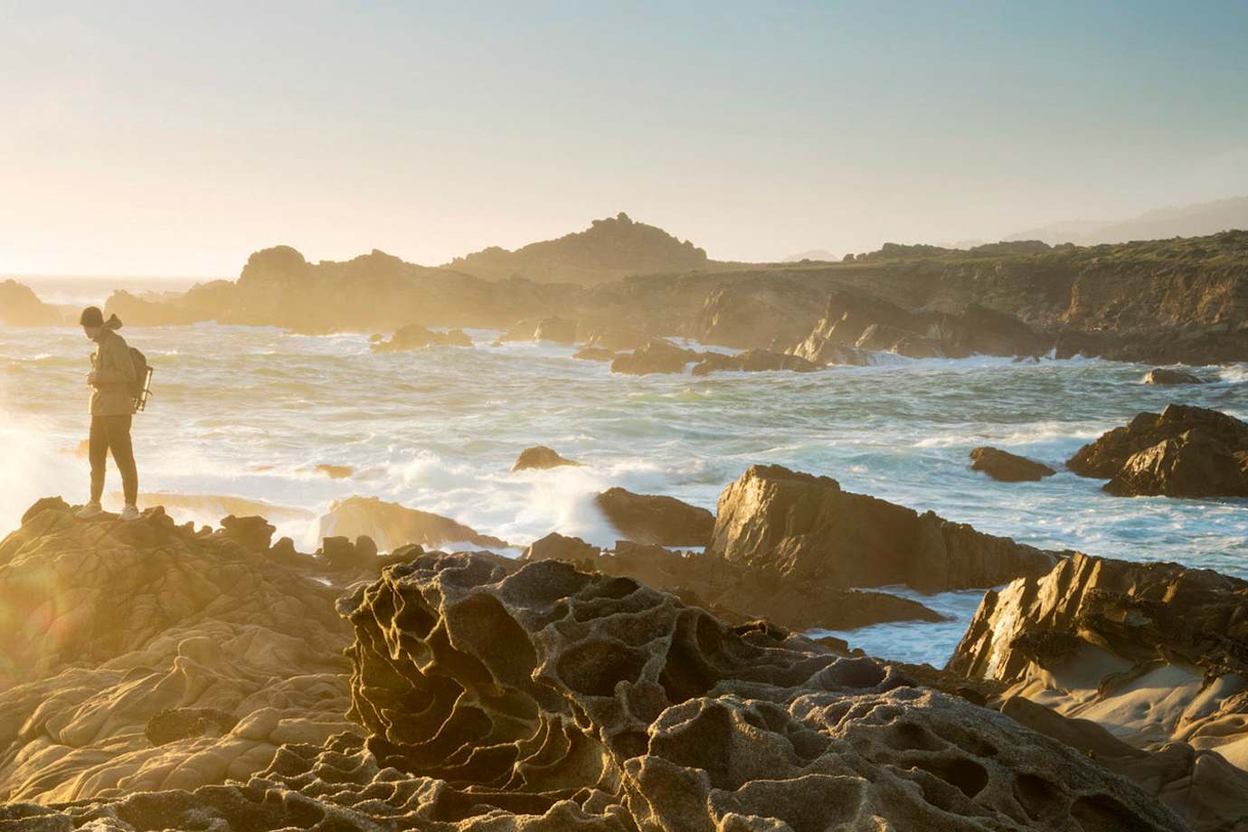 Hiker dressed warmly and standing on the honeycombed rocks known as tafoni looks out to sea at sunset at Salt Point State Park on the Sonoma Coast in California