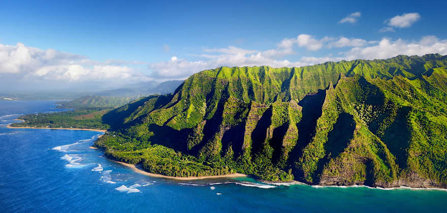 aerial view of surf breaking against rocky and lush Nāpali Coast on Kauai, Hawaii.