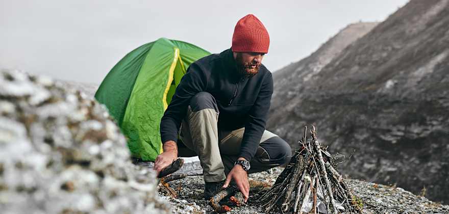 a hiker in a red beanie builds a campfire in front of a green tent