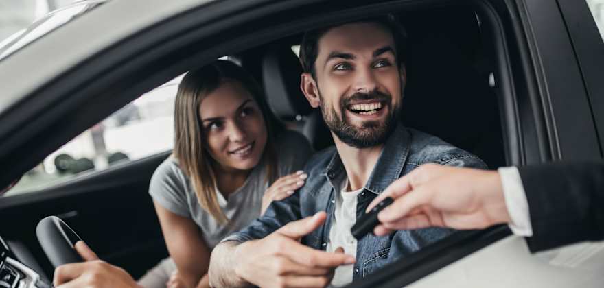 A couple smiles while sitting in their new car as they are handed the keys