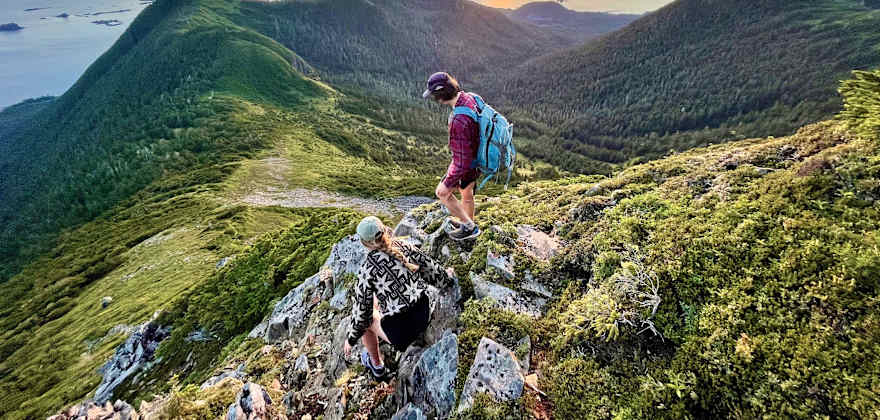 Two hikers scramble on rocks along trail overlooking Sikta sound at sunset. 