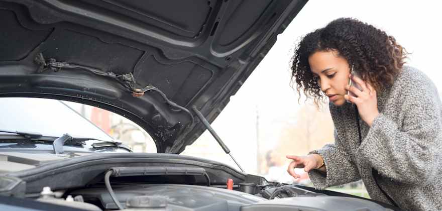 A woman looks under the hood of her car while on the phone.