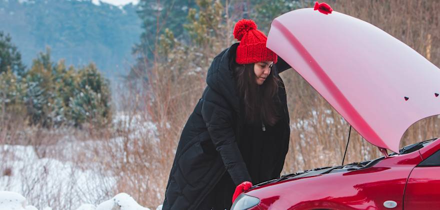 A woman in a coat and hat looks under the hood of her red car.