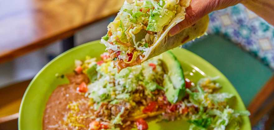 A hand holds a stuffed soft taco over a traditional Mexican combo plate. 