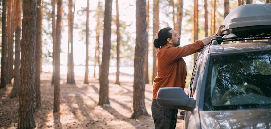 A person opens a roof-top storage box on their SUV parked in forest.