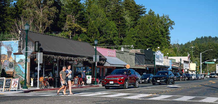 two people walk across the street in a picturesque small town with trees and mountains visible against a blue sky
