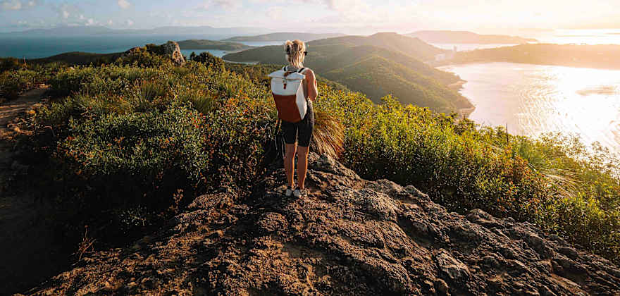 a woman searing shorts and a backpack stands on top of a mountain looking out at the ocean in the sun