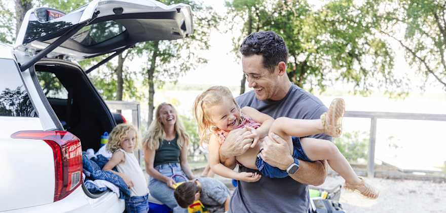 A family laughs and plays as they remove their luggage from the back of their white SUV.