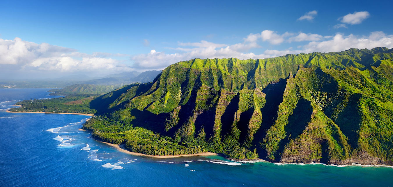 aerial view of surf breaking against rocky and lush Nāpali Coast on Kauai, Hawaii.