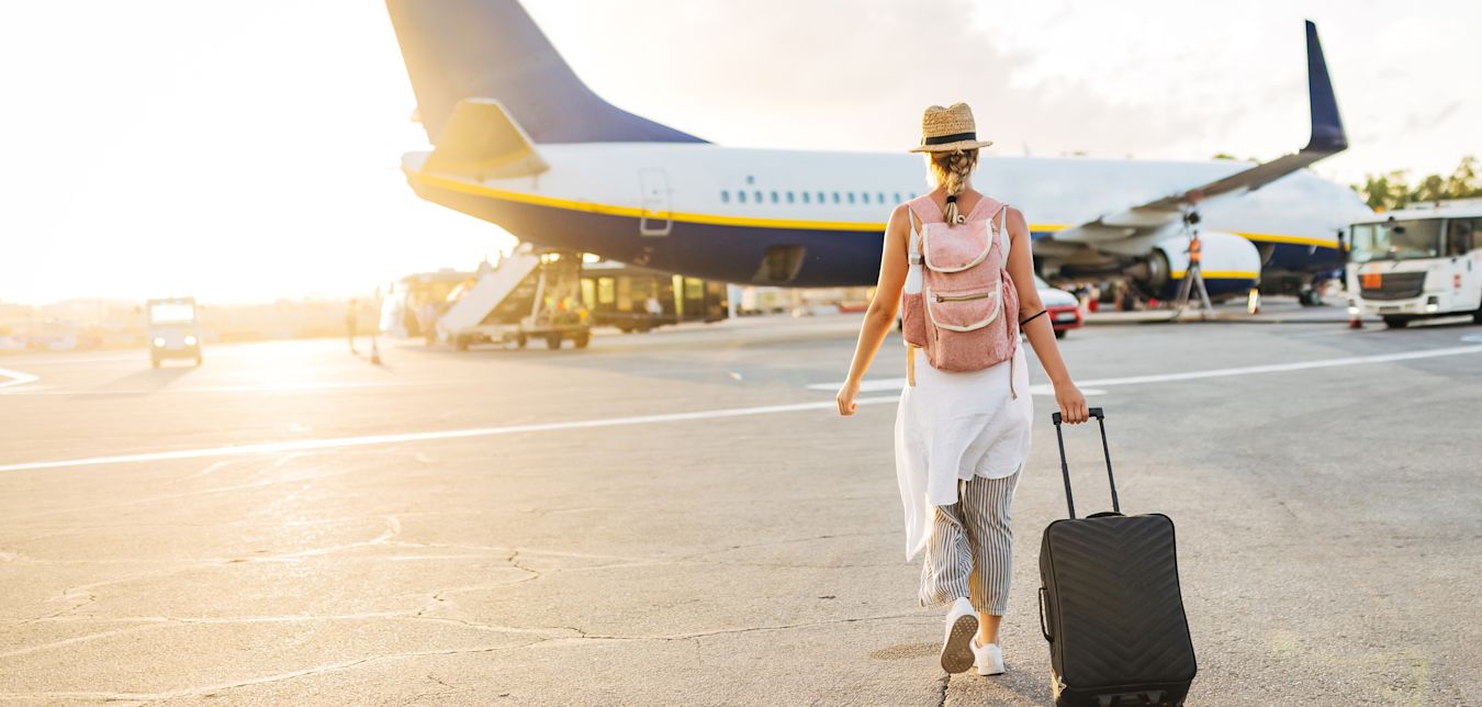 A woman rolling a suitcase walks towards an airplane as the sun sets.