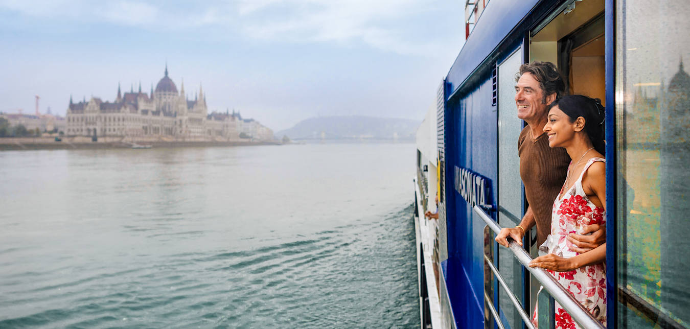 A man and woman standing on a river cruise ship balcony. The Budapest skyline is in the distance.