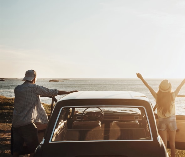 picture of a couple on a road trip standing outside their car looking at the sun setting over the ocean