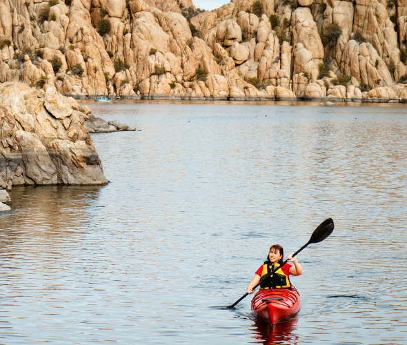 A woman paddles a kayak on Watson Lake in Prescott, Arizona with rocky walls behind her.