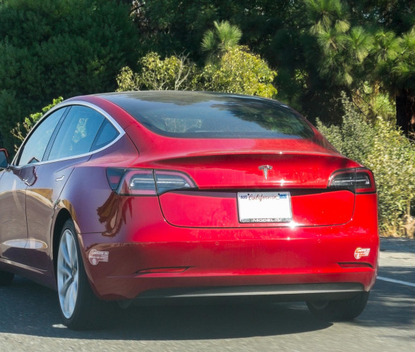 Red Tesla Model 3 driving with carpool stickers on the bumper.